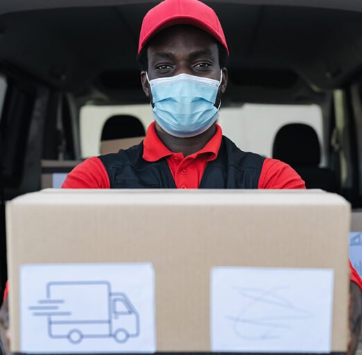 African delivery man carrying cardboard box while wearing face mask to avoid corona virus spread - People working with fast deliver during corona virus outbreak