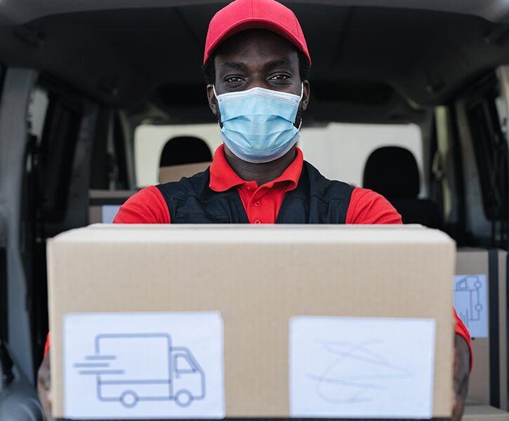 African delivery man carrying cardboard box while wearing face mask to avoid corona virus spread - People working with fast deliver during corona virus outbreak