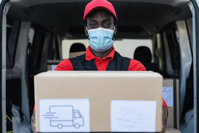 African delivery man carrying cardboard box while wearing face mask to avoid corona virus spread - People working with fast deliver during corona virus outbreak