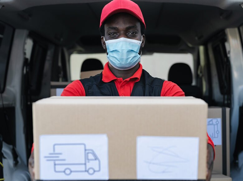 African delivery man carrying cardboard box while wearing face mask to avoid corona virus spread - People working with fast deliver during corona virus outbreak
