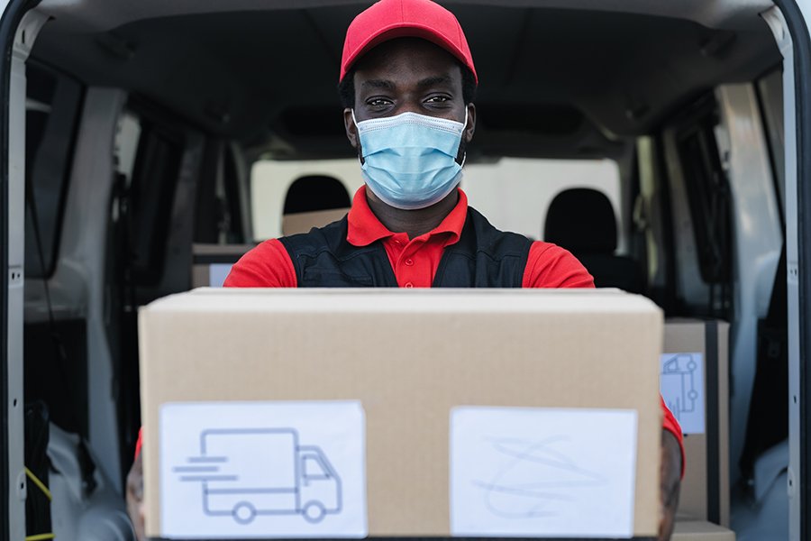African delivery man carrying cardboard box while wearing face mask to avoid corona virus spread - People working with fast deliver during corona virus outbreak